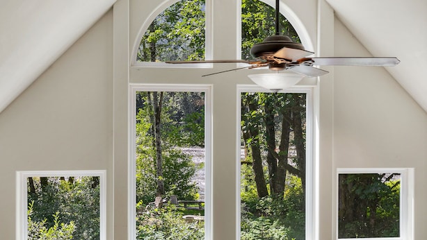 A room with a sloped ceiling, a ceiling fan, and four windows offering views of a wooded area.