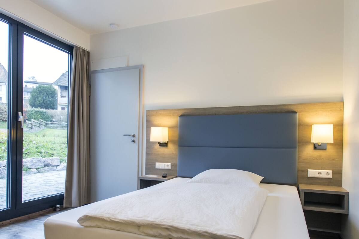 Ein modernes Hotelzimmer mit einem großen Bett, Holzboden und Blick durch eine Glastür.