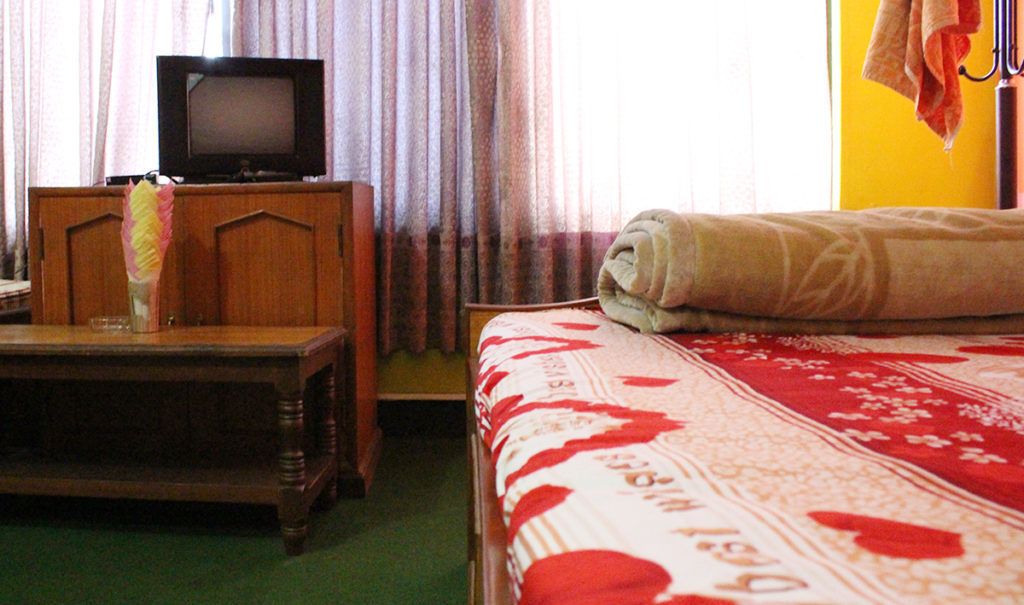 Una habitación con una cama, una mesita de noche de madera, un televisor sobre un mueble y una ventana con cortinas.