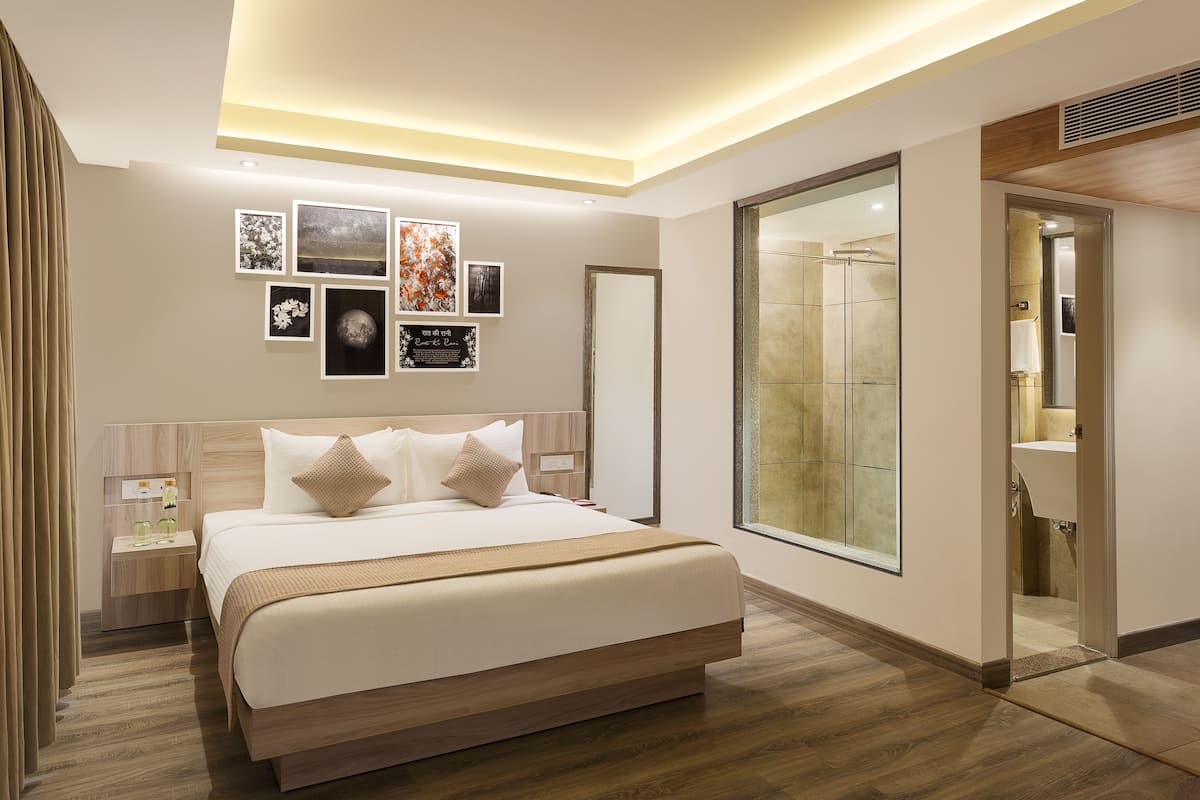Una habitación de hotel moderna con una cama grande, una mesita de noche, un televisor montado en la pared y un baño visible a través de una puerta de cristal.