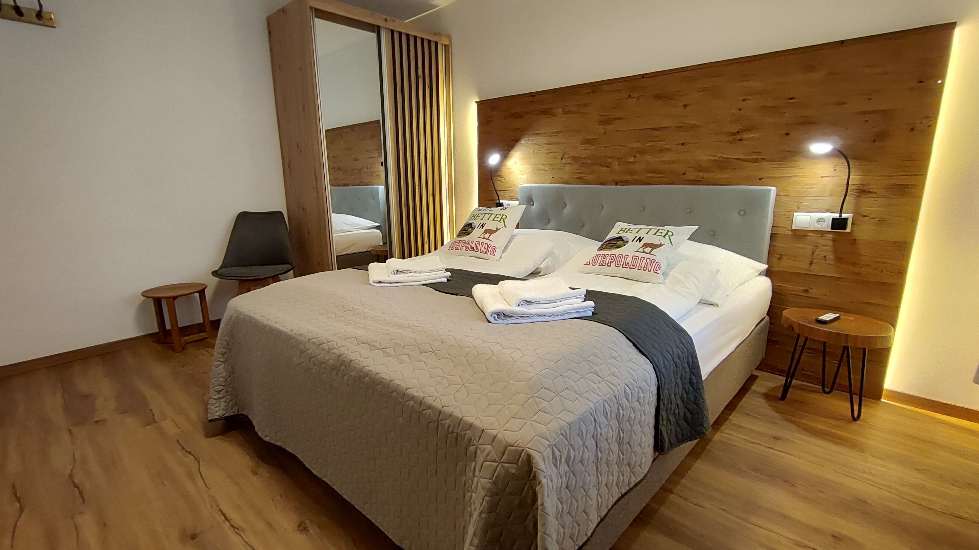 A neatly made bed with a quilted bedspread, two pillows with a 'Better in Ultrapolding' logo, a bedside table with a lamp, and a chair in a room with wooden flooring and a wooden headboard.