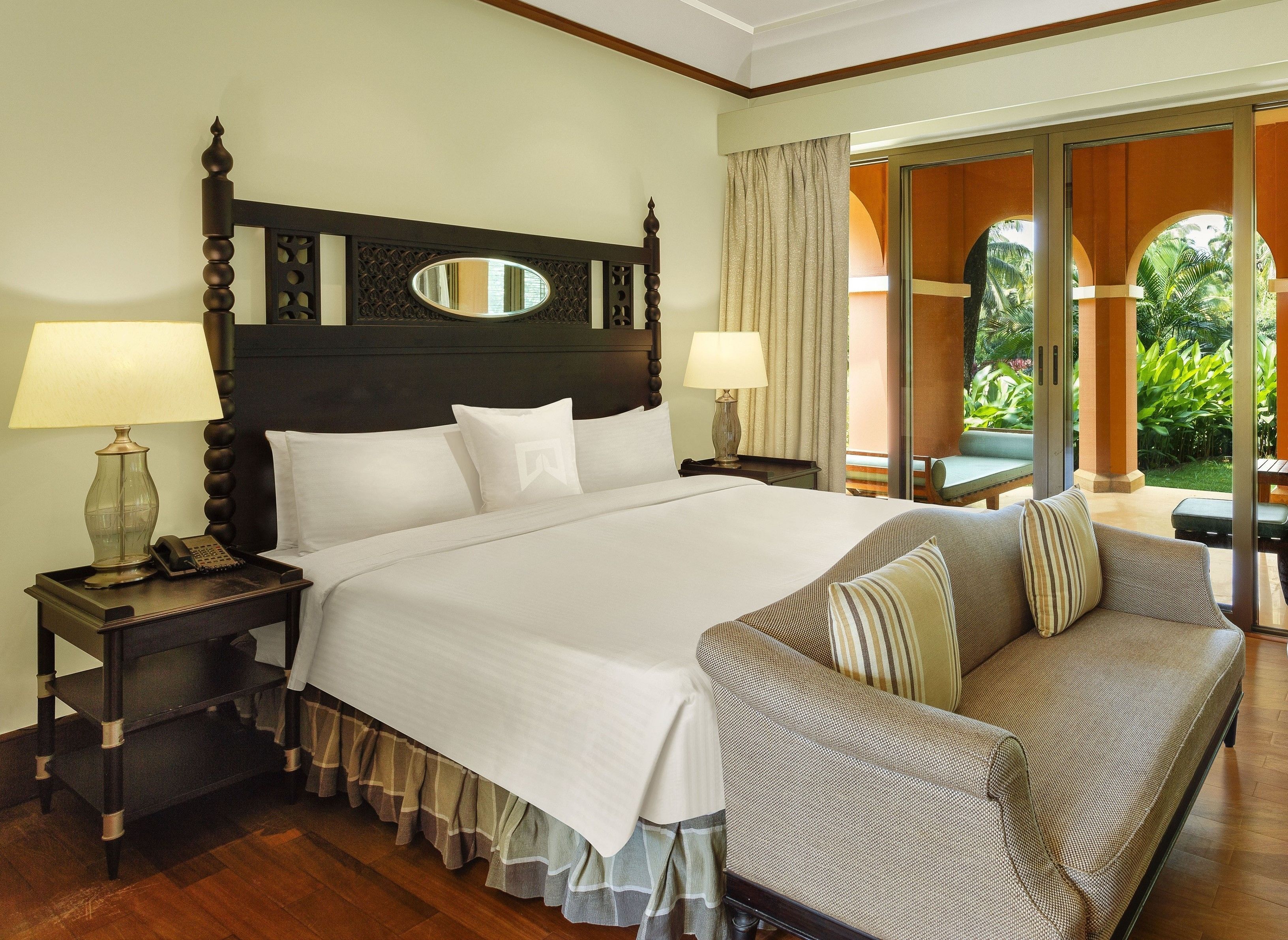 A hotel room with a large bed, two bedside tables with lamps, a sofa, and a view of greenery through a glass door.