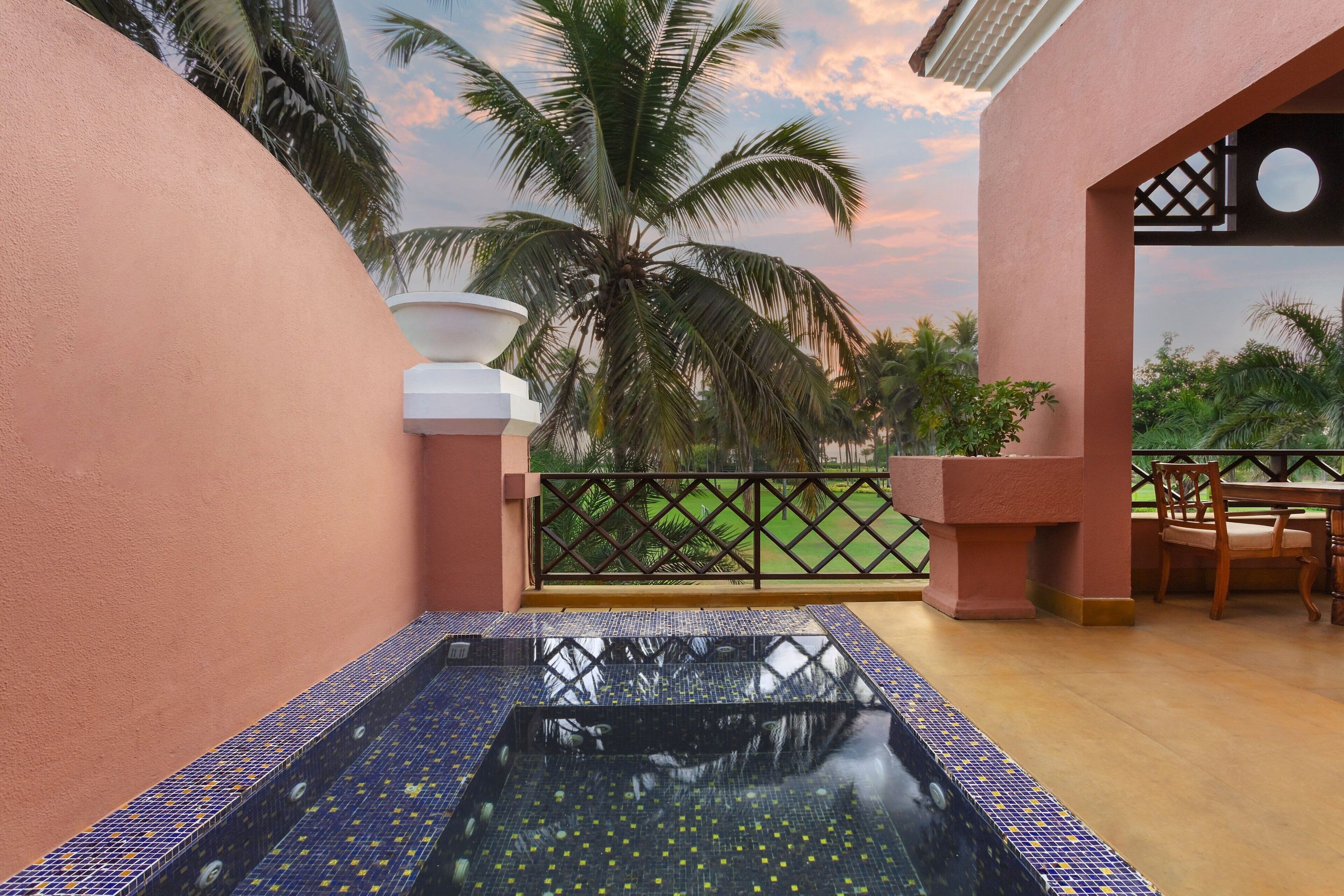 Un área de spa con un jacuzzi de azulejos, una mesa de madera con sillas y vistas a palmeras y un atardecer.