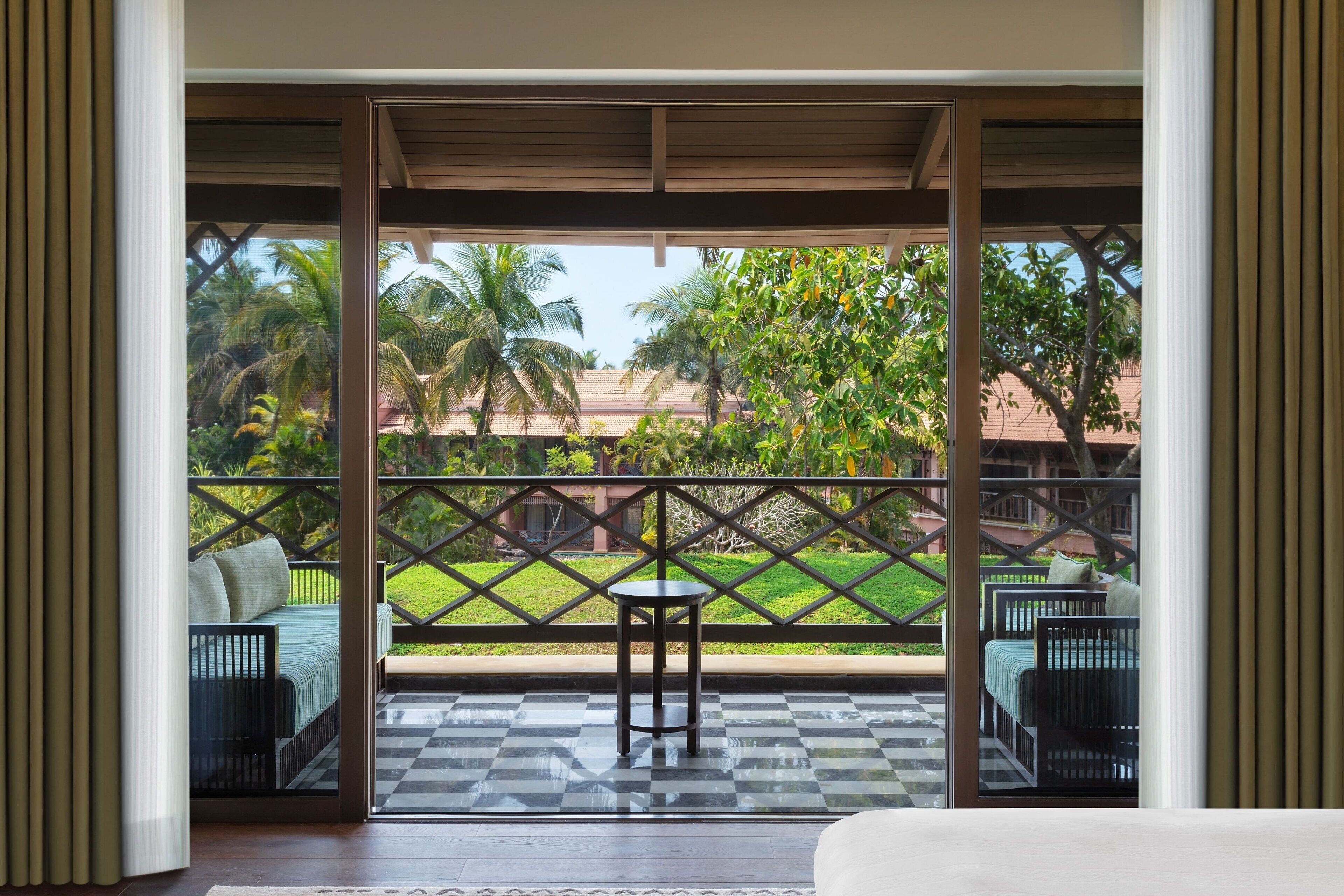 A room with a checkered floor, a bed, a table, and chairs. There is a view of a garden and palm trees outside.