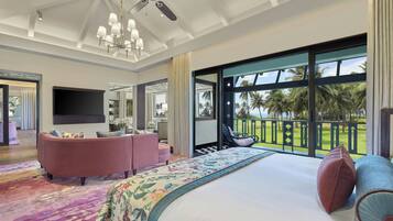 A spacious bedroom with a large bed, a seating area with a sofa and armchairs, a television, and a balcony with a view of palm trees.