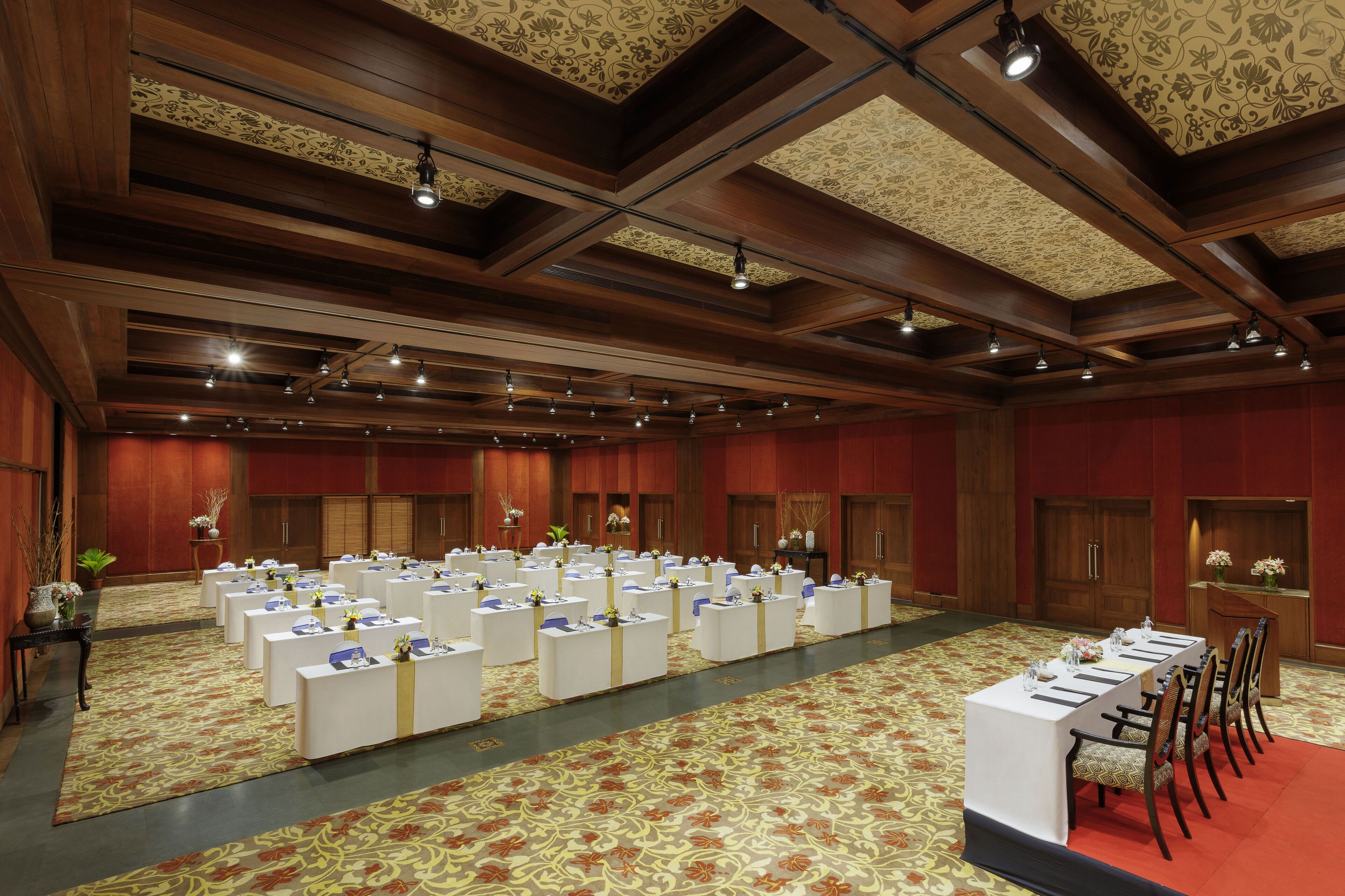 A large conference room with rows of white tables and chairs, wooden ceiling beams, and patterned carpet.
