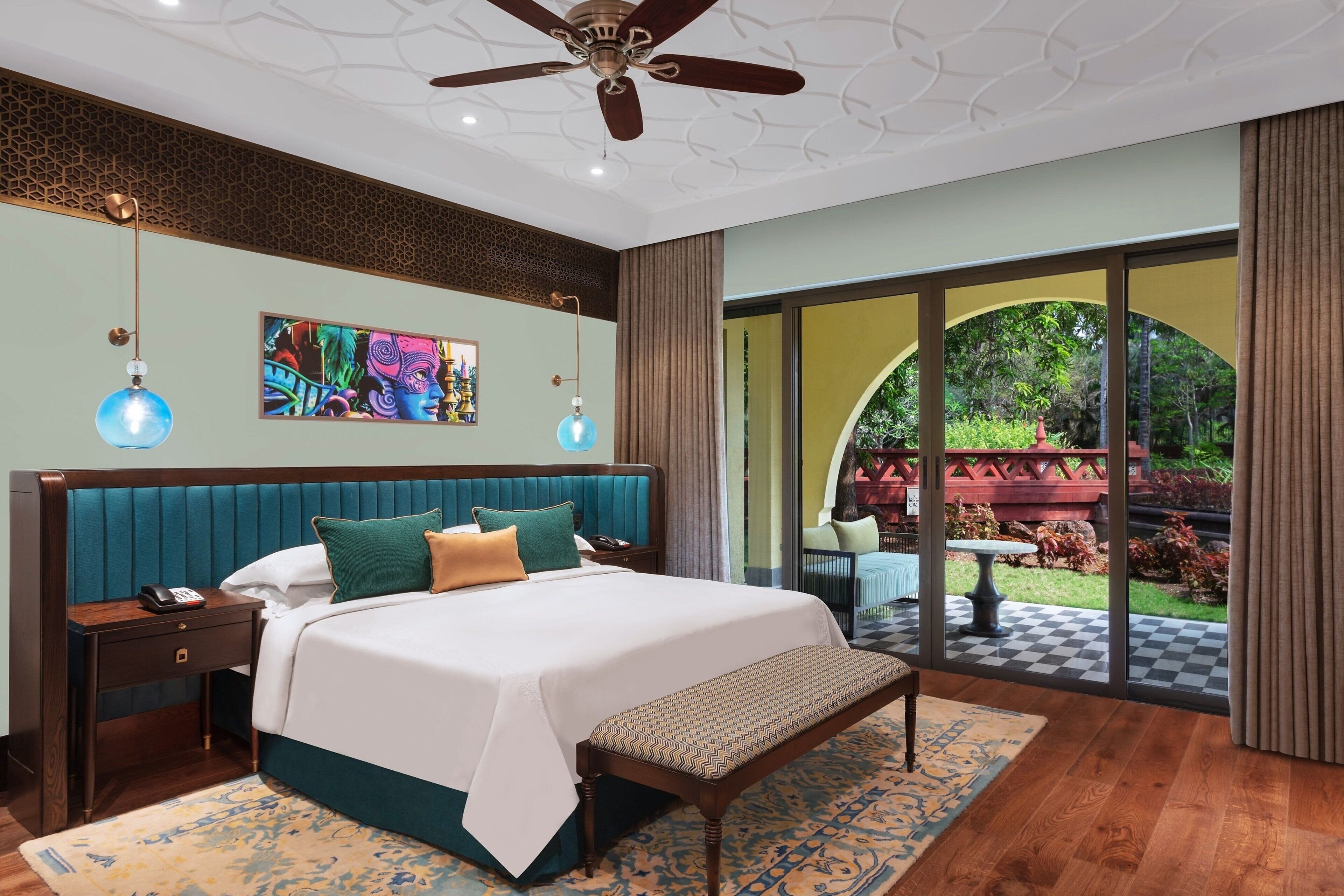 A bedroom with a large bed, a bench, a nightstand, a ceiling fan, and a view of the garden through a glass door.