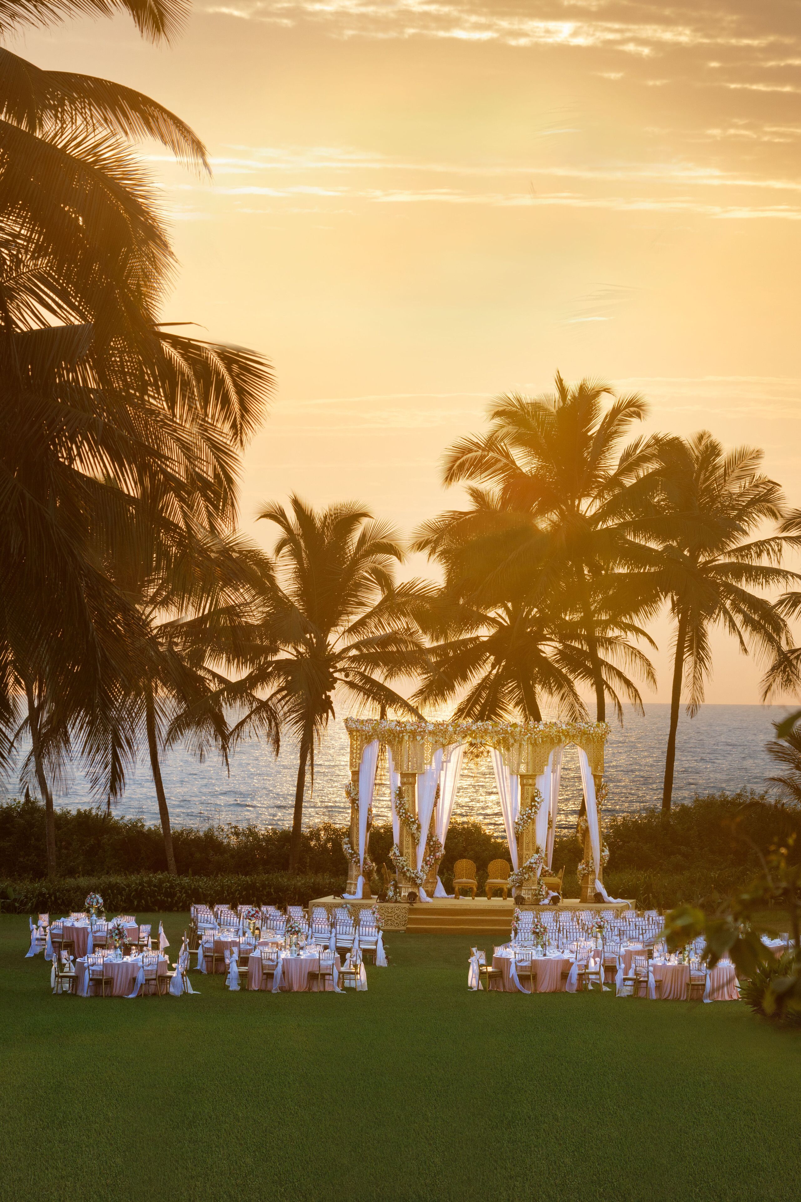 A wedding ceremony setup with tables and chairs arranged for guests, palm trees, and a view of the ocean.