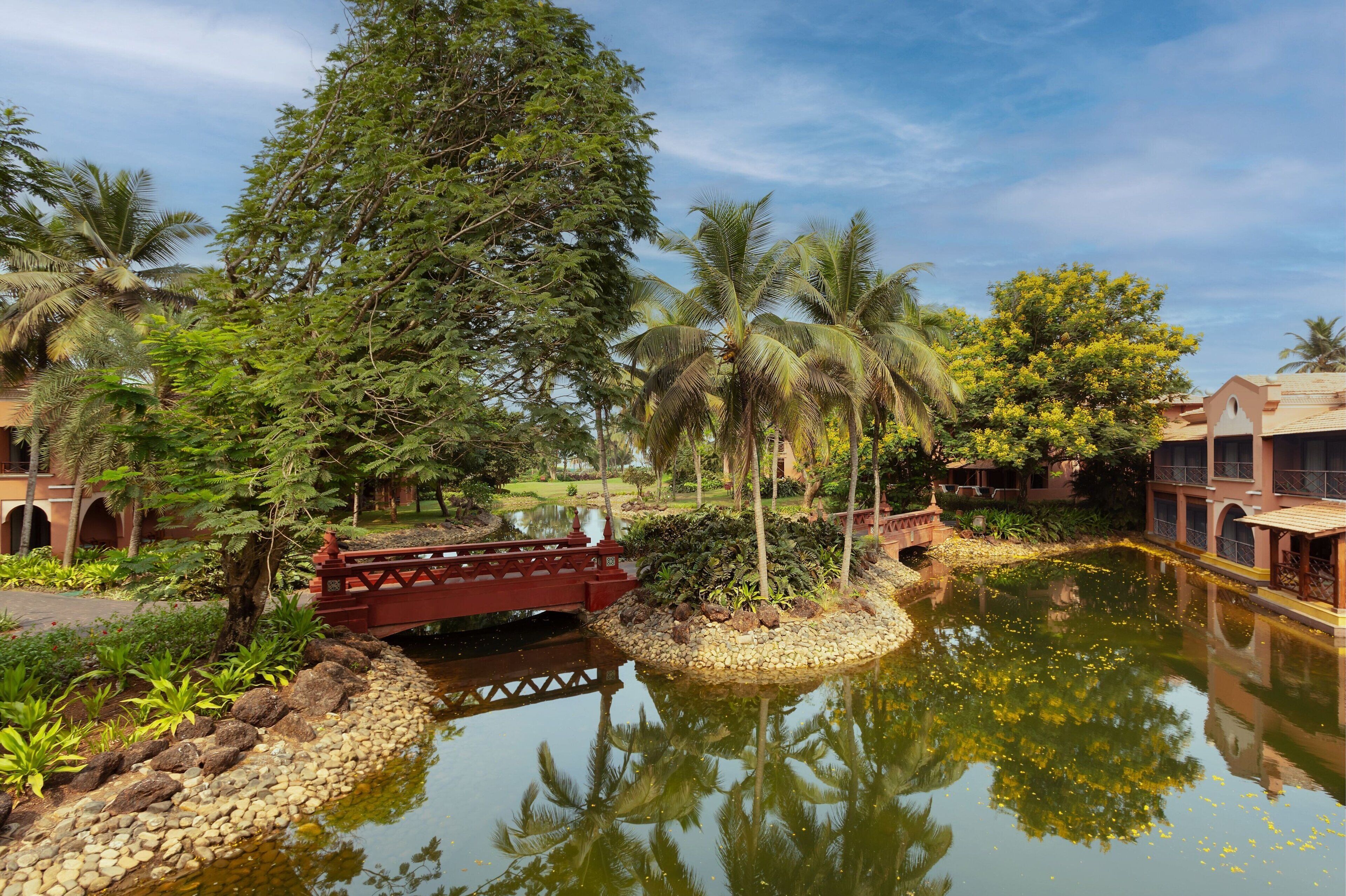 A serene water feature with a red bridge, surrounded by lush greenery and palm trees, reflecting the sky and nearby buildings.