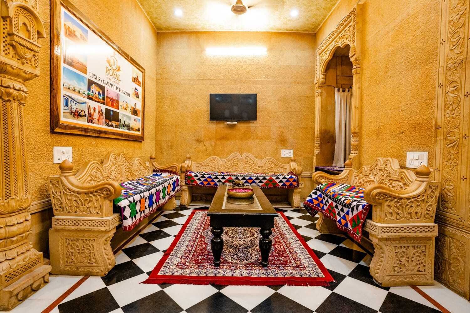 Een kamer met een geruit vloertje, twee houten banken met kleurrijke dekens, een centrale tafel en een televisie aan de muur.