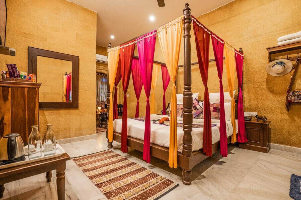 Un lit  à baldaquin avec des rideaux rouges et orange, une table de chevet en bois, un miroir et un tapis à motifs.