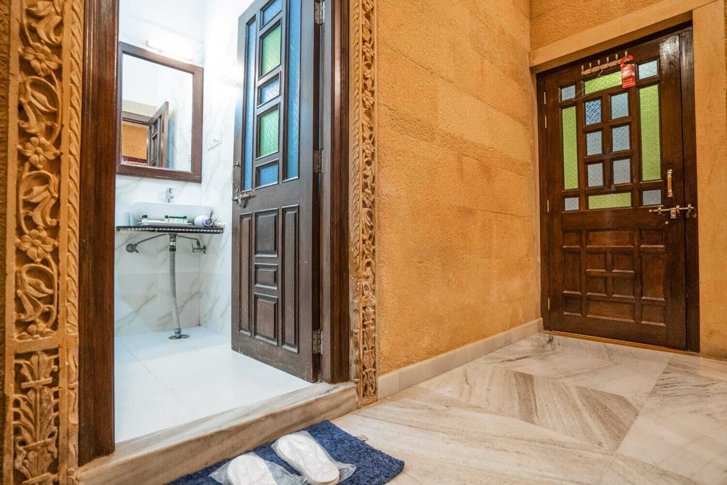 Un couloir doté d’un sol en marbre, une porte en bois avec un panneau de verre vert, et une salle de bain visible à travers une porte entrouverte.