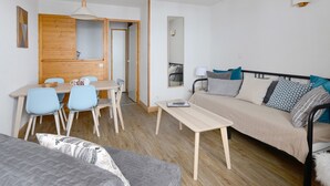 Een knus woongedeelte met een houten eettafel, blauwe stoelen, een bank met opvallende kussens en een klein nachtkastje.