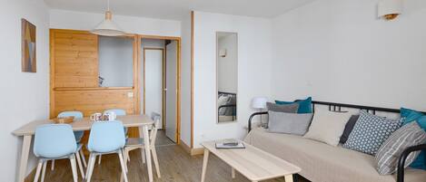 A compact living space with a wooden dining table, blue chairs, a sofa with patterned cushions, and a small bedside table.