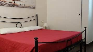 Un letto singolo con una coperta rossa in una stanza dalle pareti bianche e pavimento piastrellato.