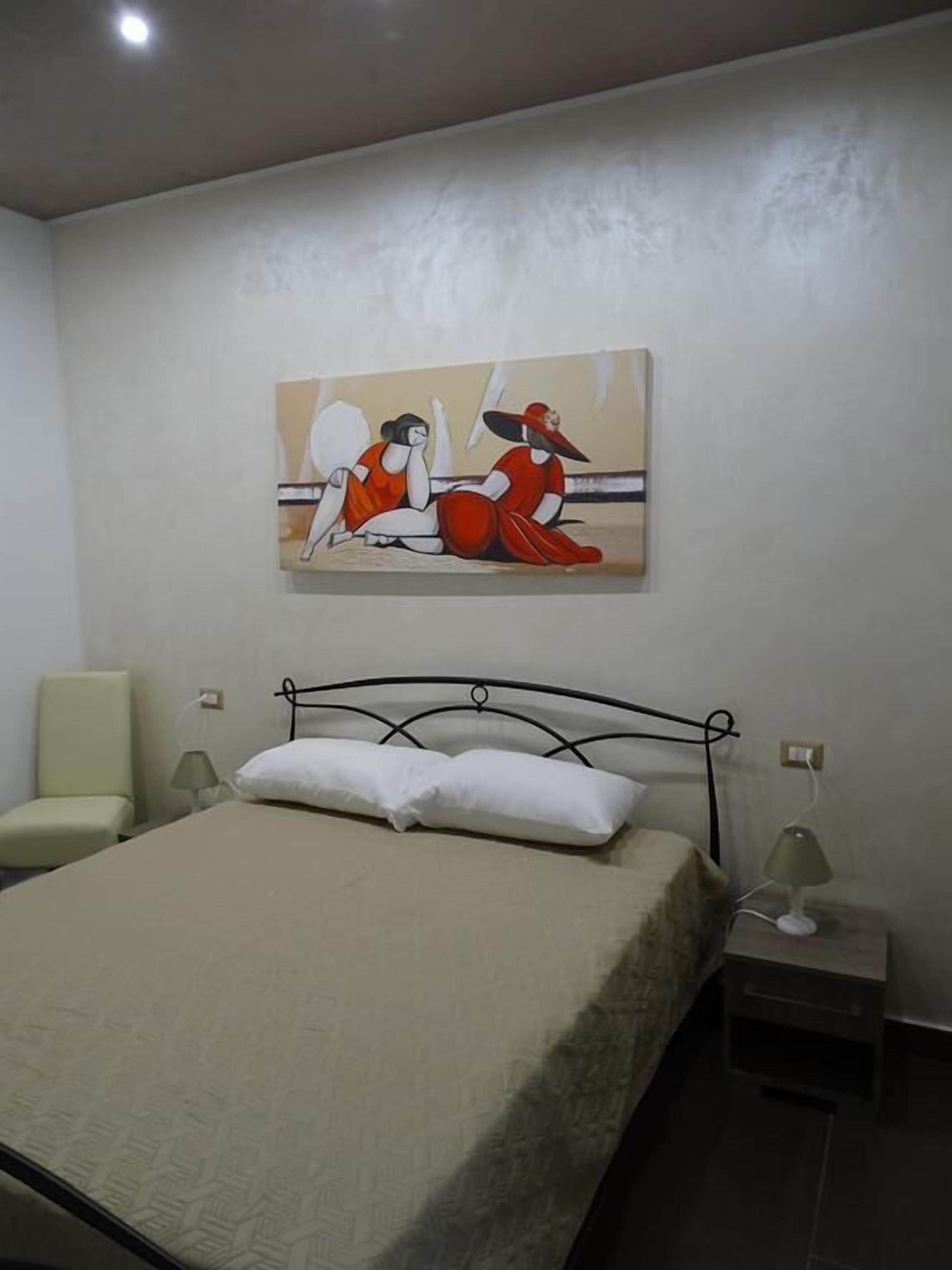 A hotel room with a bed, a chair, a nightstand with a lamp, and a painting on the wall.