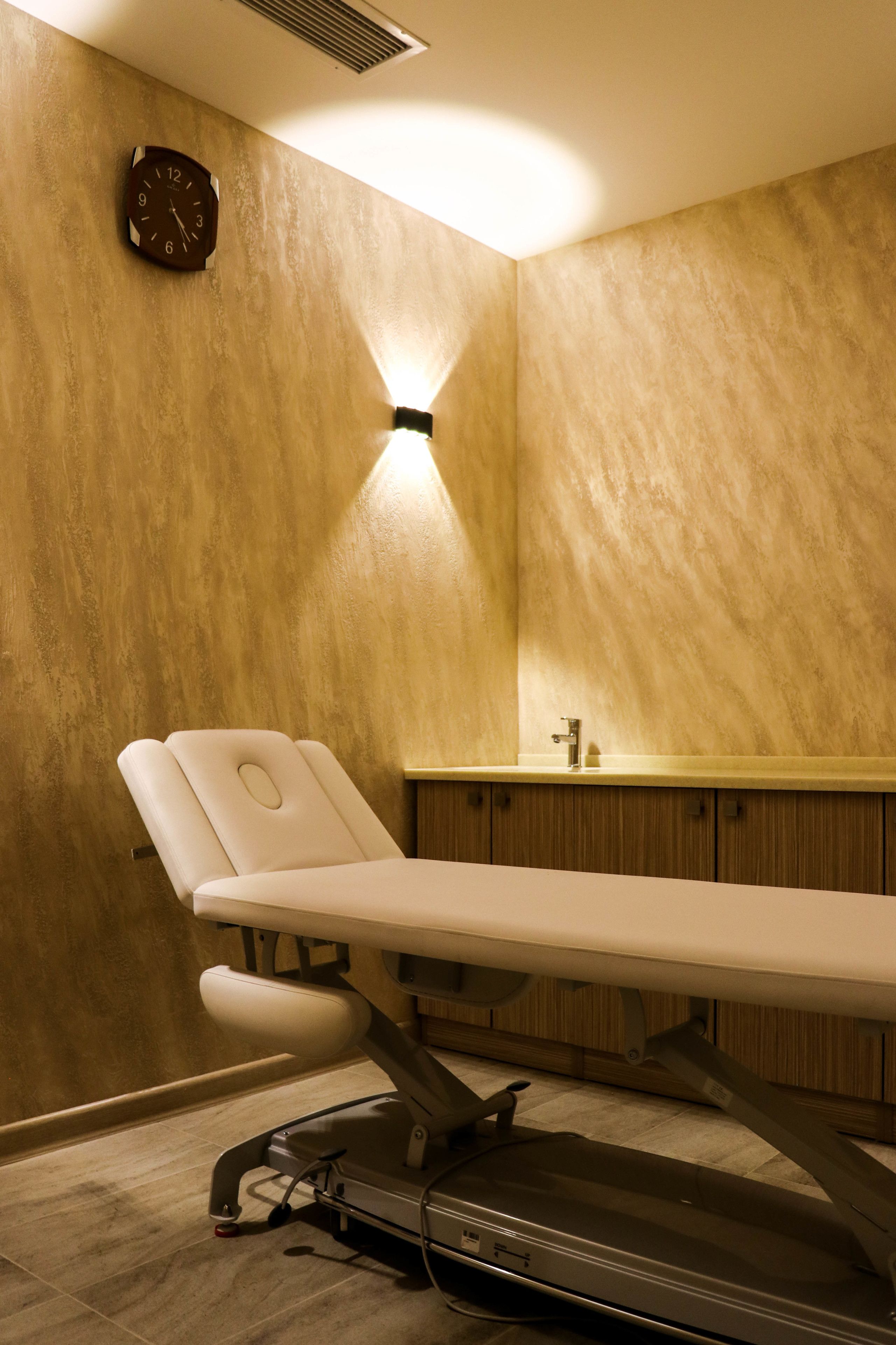 A medical examination room with a treatment table, a clock on the wall, and a sink in the background.