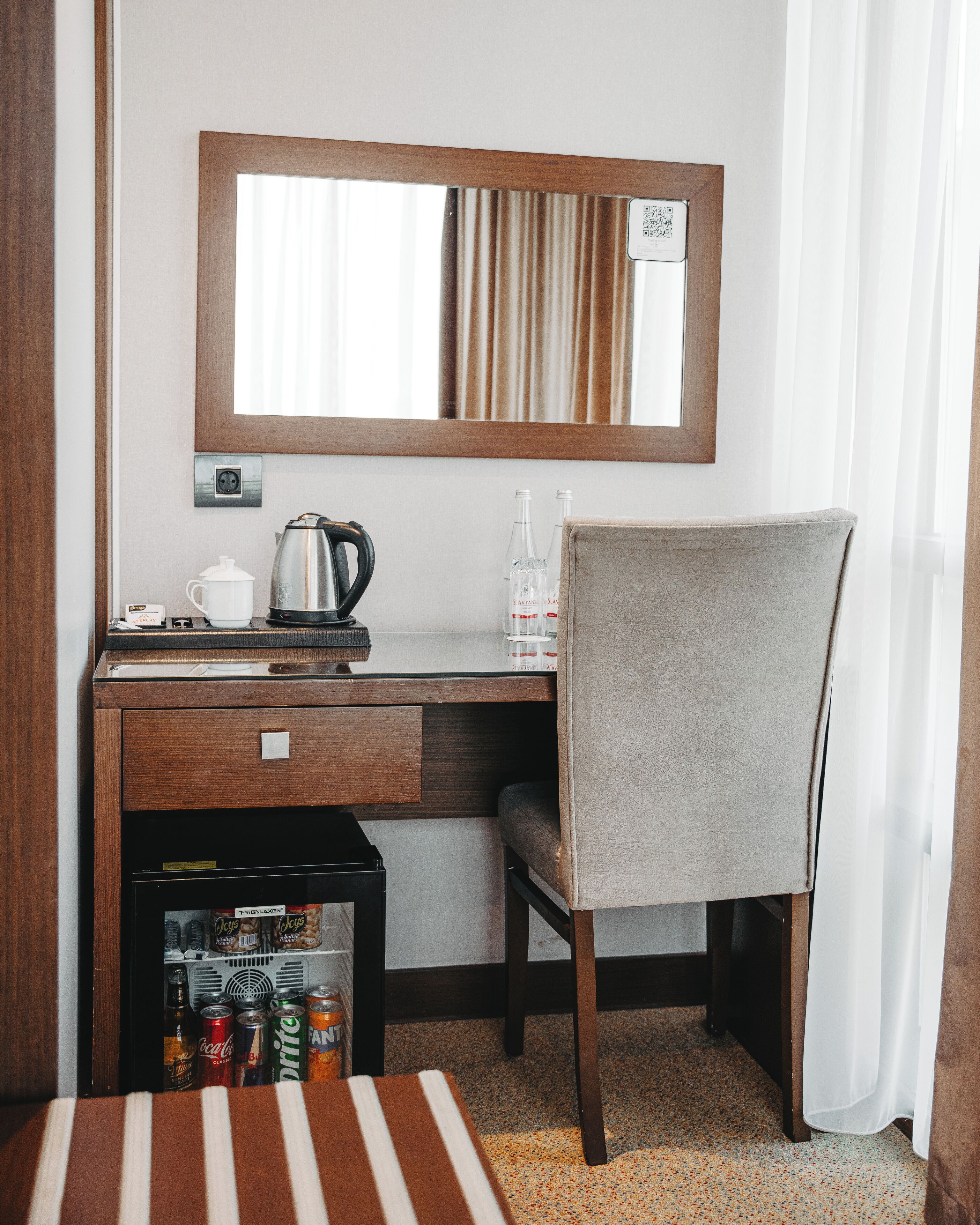 Habitación de hotel con escritorio de madera, una silla, un minibar, un espejo y una ventana con cortinas.