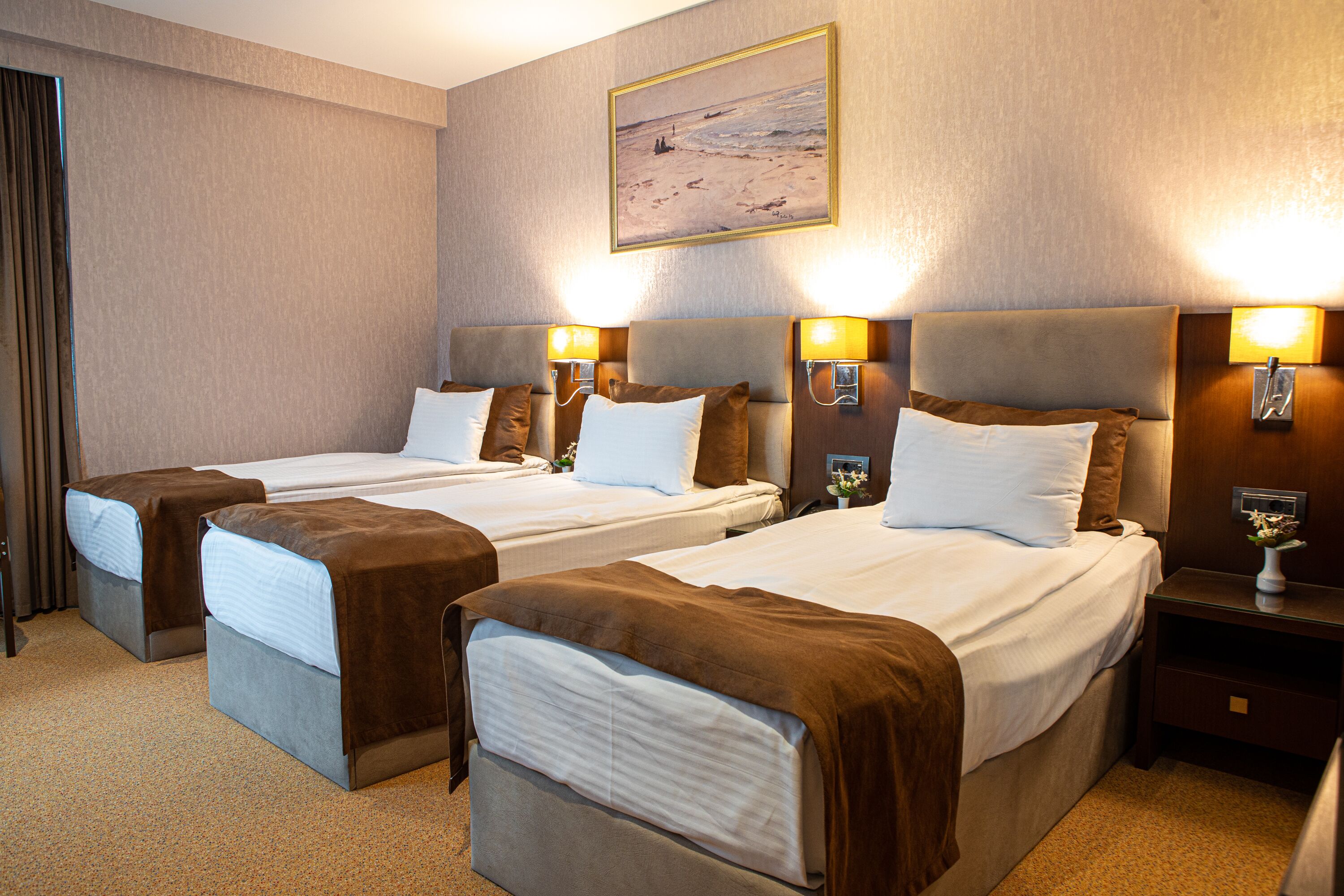 A hotel room with three single beds, each with a brown headboard and white bedding. There are two bedside tables with lamps, and a painting of a beach scene on the wall.