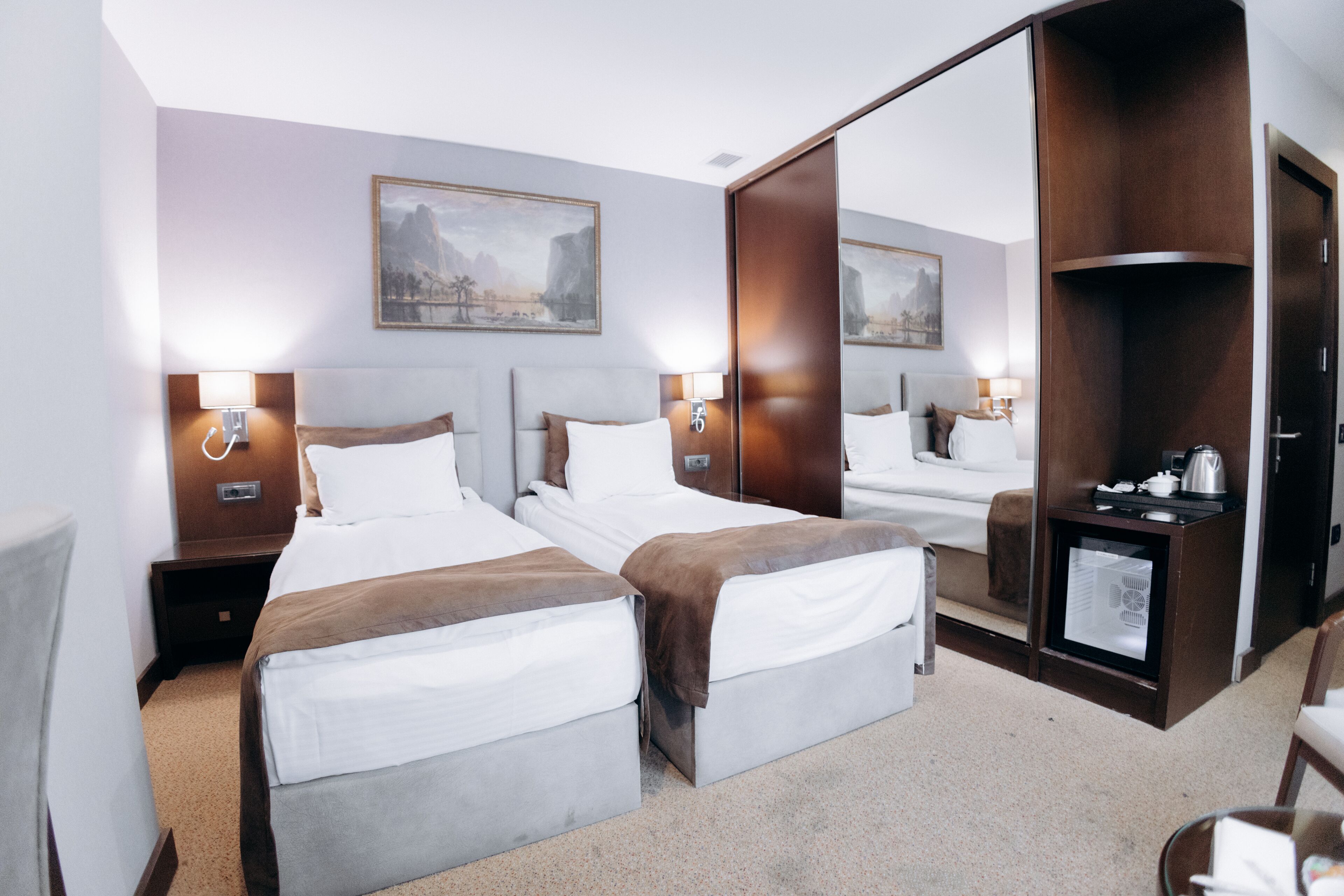A hotel room with two beds, a wardrobe, a desk with a kettle, and a small refrigerator.