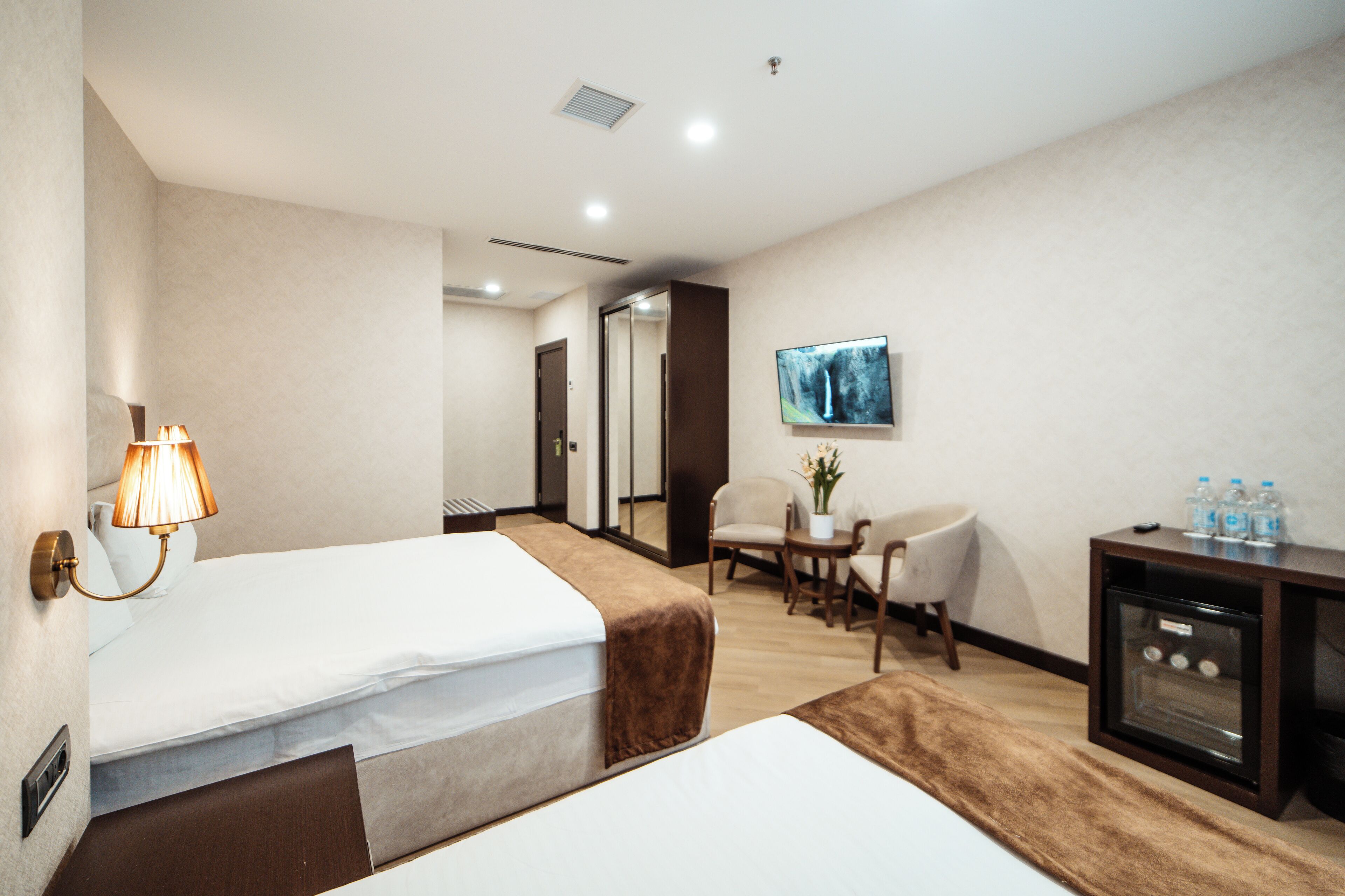 A hotel room with two beds, a desk with a TV, a chair, a small table with a plant, and a minibar.