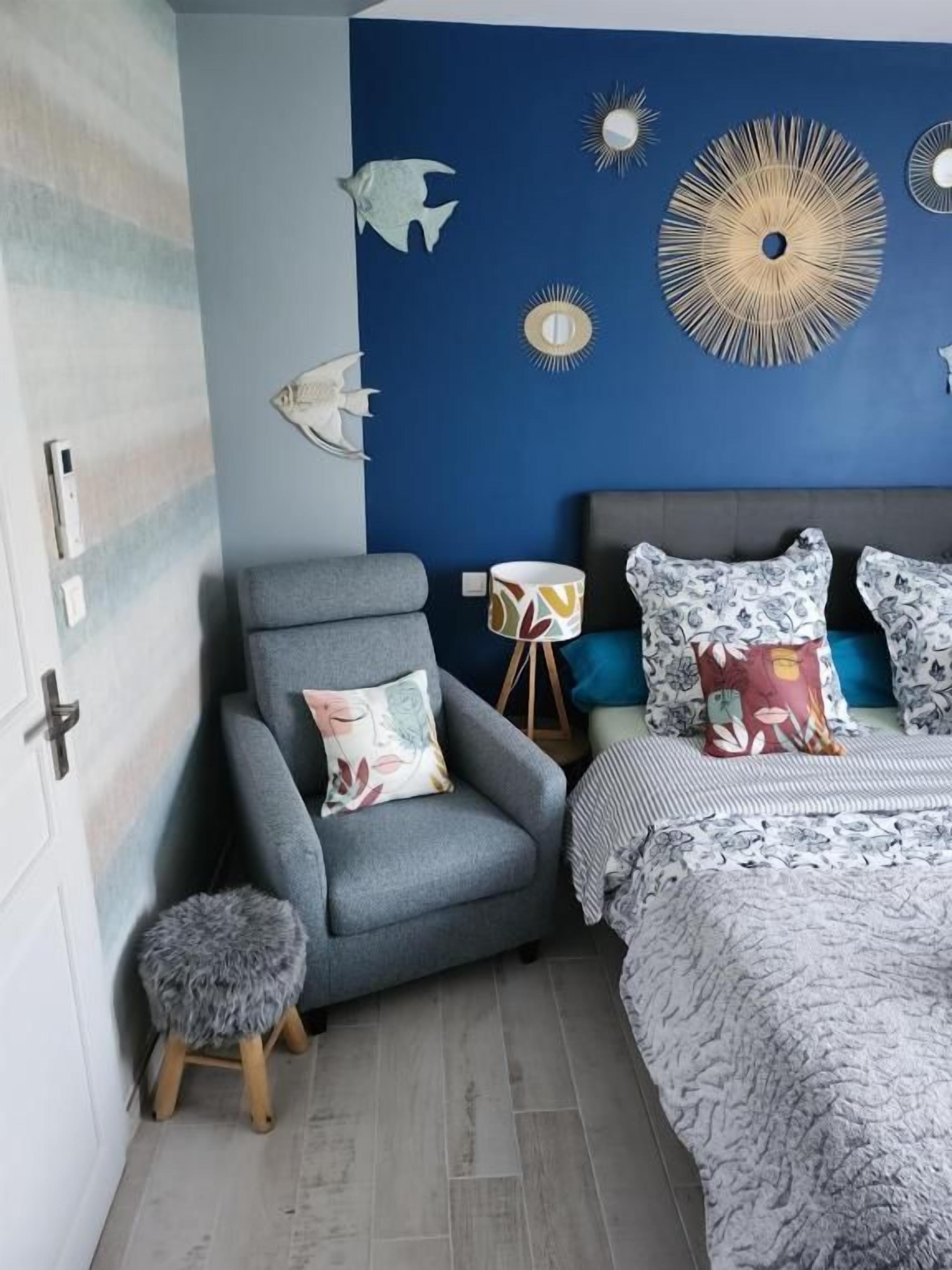 A bedroom with a blue accent wall, a grey armchair, a small stool, a bed with patterned bedding, and decorative wall art including fish and sunburst designs.