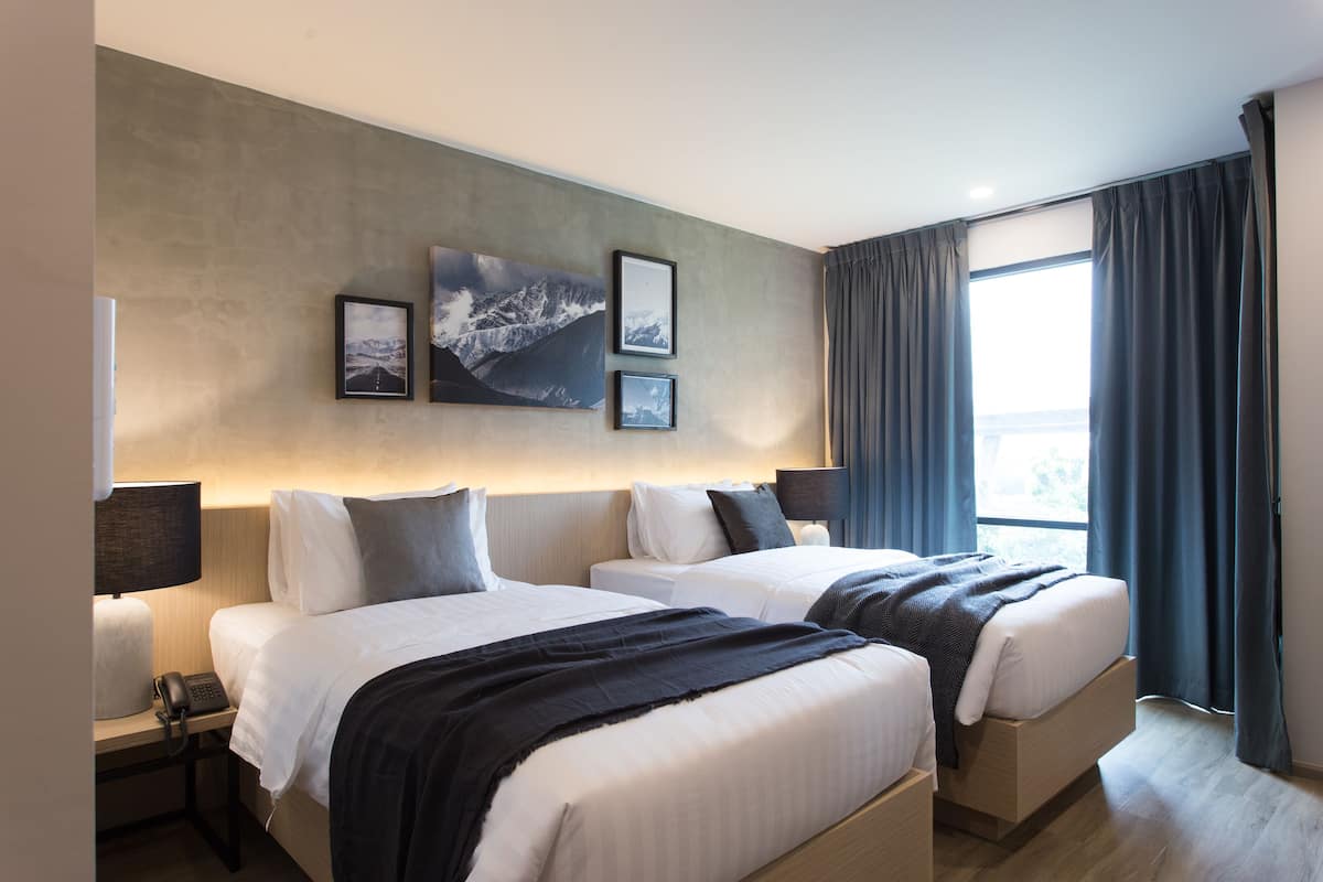 Une chambre d’hôtel avec deux lits, une grande fenêtre avec des rideaux, un téléphone fixé au mur et des tableaux encadrés.