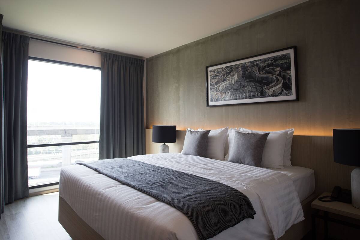 Une chambre d’hôtel avec un grand lit, une vue sur la ville, un tableau encadré au mur et une table de chevet avec une lampe.