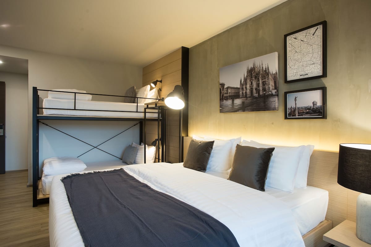 Une chambre d’hôtel avec un lit superposé, un grand lit et deux tableaux encadrés représentant Milan.