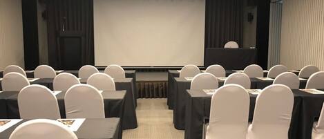 A conference room with rows of tables and chairs, a stage, and a large screen.