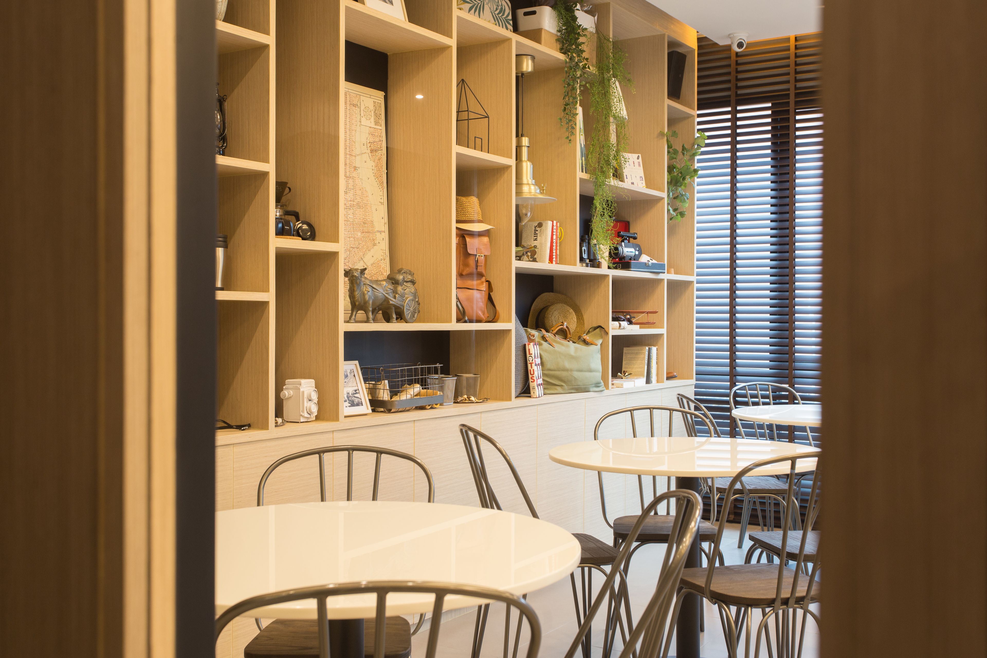 Un comedor con mesas y sillas redondas, estantes de madera con diversos objetos y una ventana con persianas.