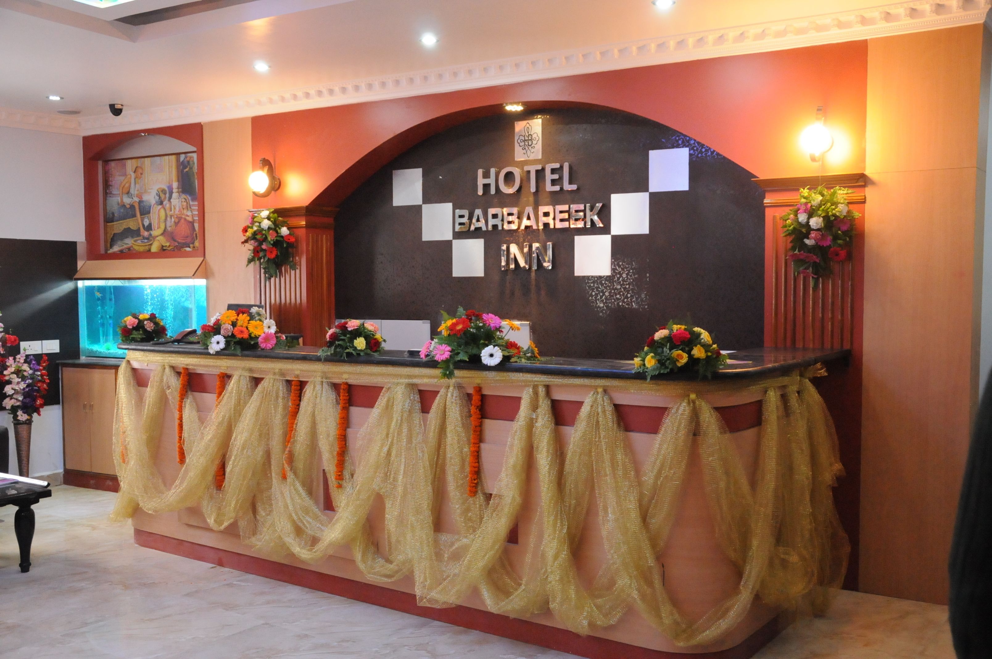A reception desk at Hotel Barbareek Inn with a dark backdrop, floral arrangements, and a sign above.