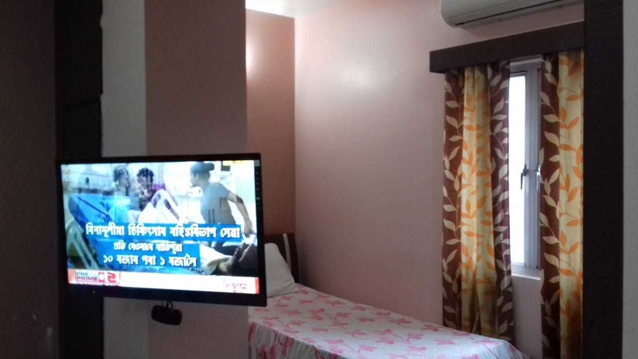 A television mounted on a wall displaying a news broadcast. There is a bed with a floral bedspread and a window with curtains.
