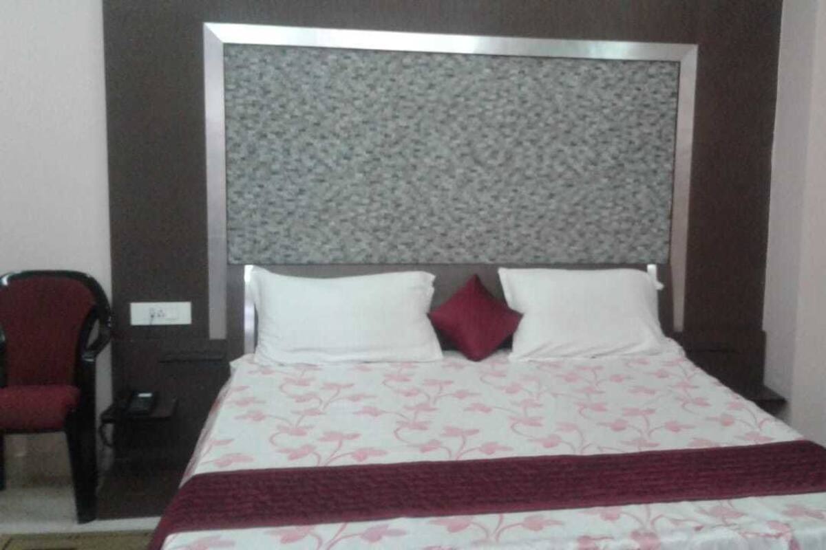 Uma cama bem arrumada com edredom floral e duas almofadas, uma cadeira vermelha e uma pequena mesa de cabeceira.