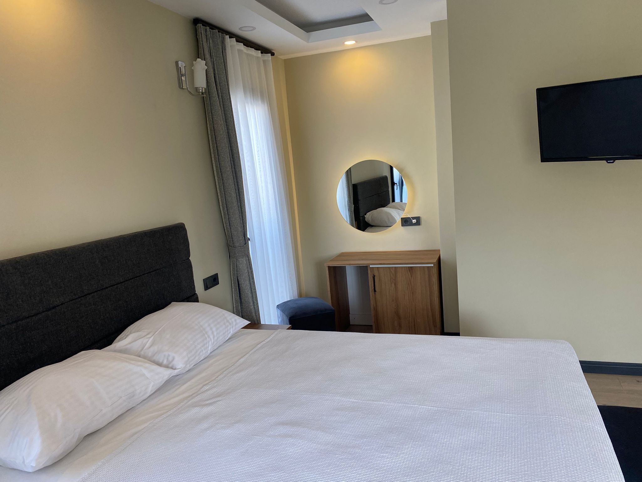 Habitación de hotel con una cama grande, una mesita de noche, un espejo y un televisor montado en la pared.