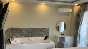 A modern hotel room with a large bathtub, a bedside table, a bench, a mirror, and an air conditioning unit.