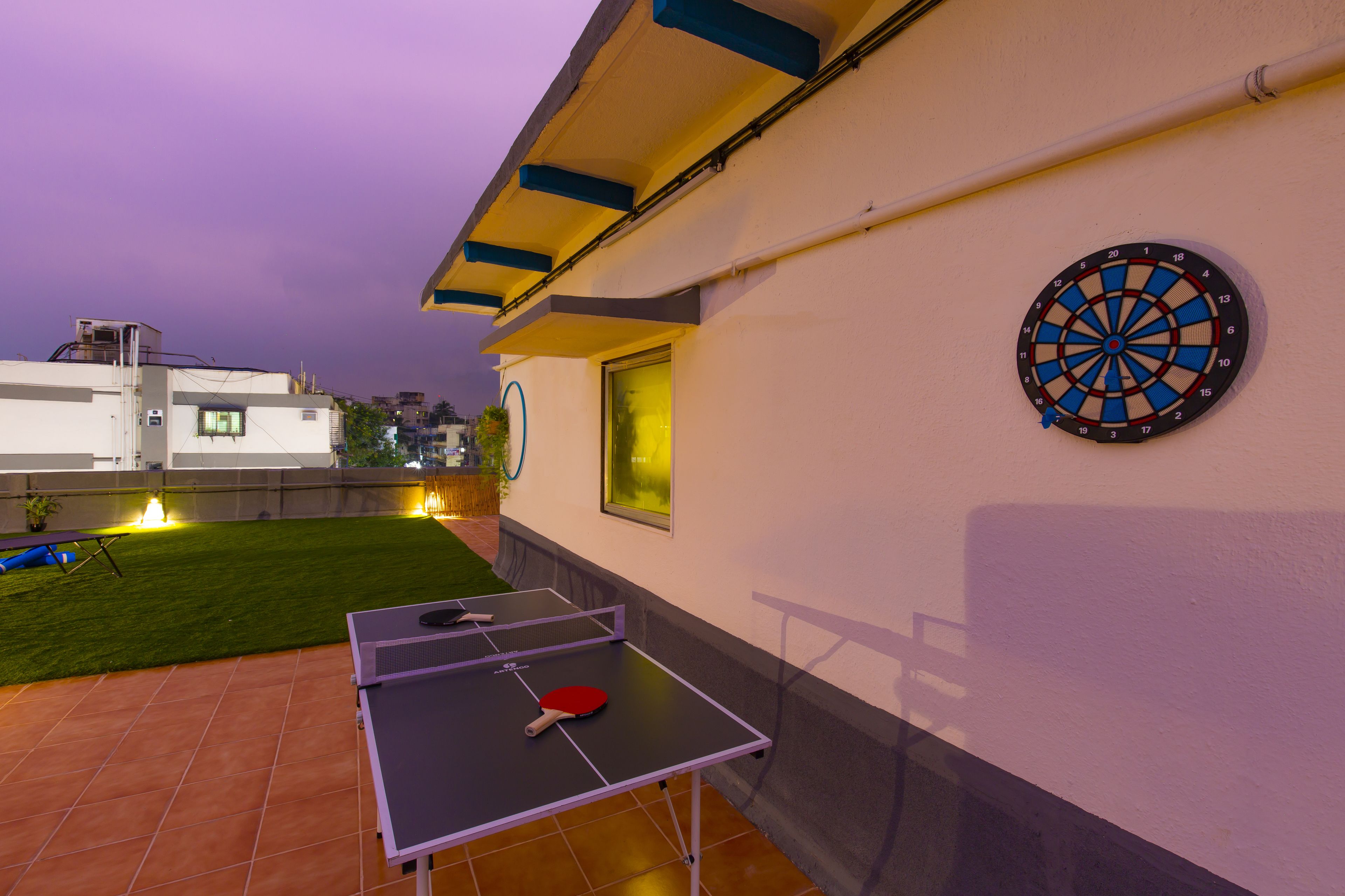 Une table de ping-pong et un jeu de fléchettes sont installés sur une terrasse, avec une pelouse verdoyante et un bâtiment en arrière-plan.