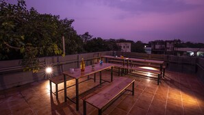 An outdoor terrace with wooden tables and benches, surrounded by a low wall and trees. - Locomo Hostel OG - Stay Work Eat (Mumbai)