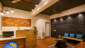 A modern reception area with a wooden desk, a water dispenser, a seating area with blue cushions, and a blackboard with maps and information. - Locomo Hostel OG - Stay Work Eat (Mumbai)