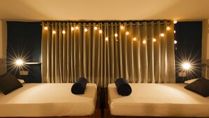 A hotel room with two beds, a curtain with string lights, and a nightstand with a lamp. - Locomo Hostel OG - Stay Work Eat (Mumbai)