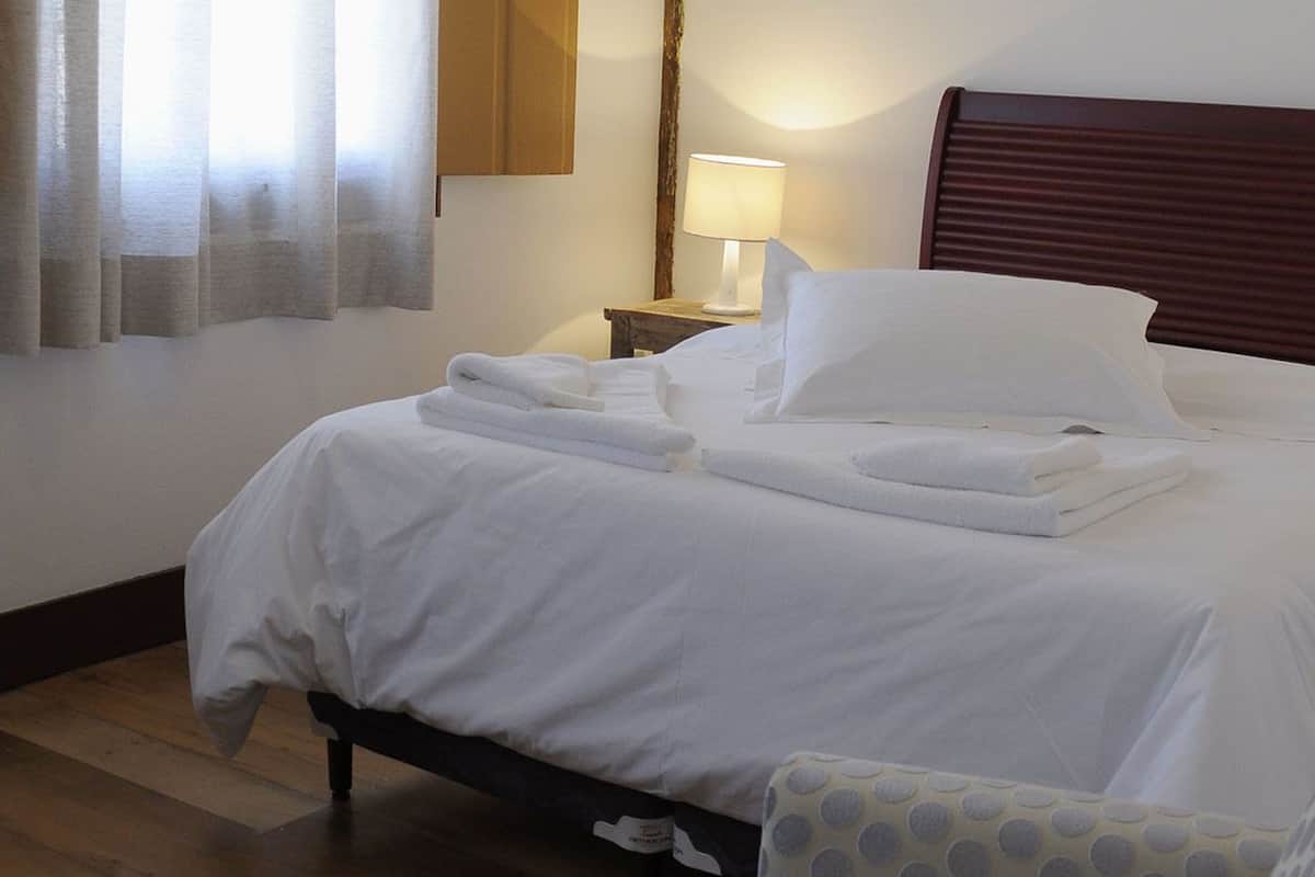 A neatly made bed with white linens and towels, a wooden headboard, a bedside lamp, and a chair with a polka dot cushion.