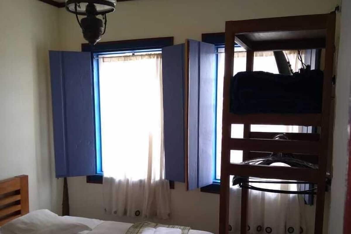 A room with a bed, a window with blue shutters, a wooden ladder, and a hanging light fixture.