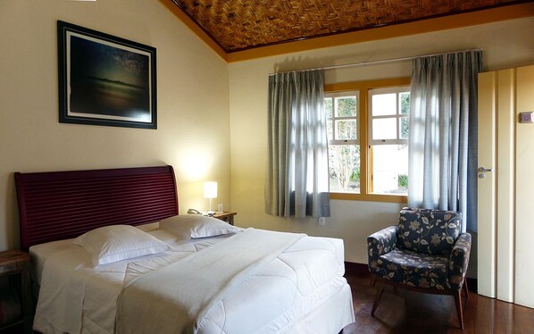 A hotel room with a bed, a chair, a nightstand, a lamp, a window with curtains, and a framed picture on the wall.