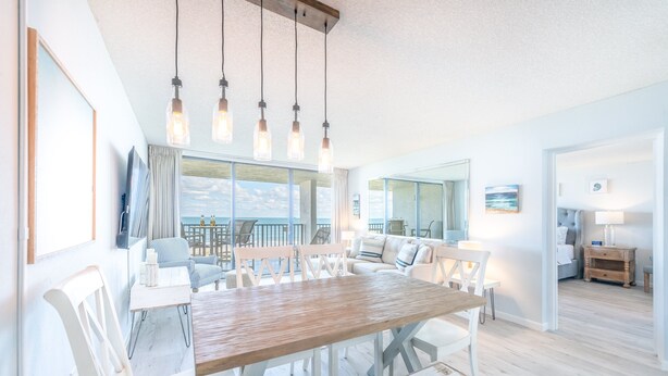 A spacious dining area with a large wooden table and white chairs, a television, and a view of the ocean.