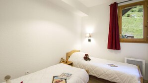 A bedroom with two beds, a window with a red curtain, a wall-mounted lamp, and a view of a green landscape. - Chalet in French Alps with Scenic Views (VALMEINIER)