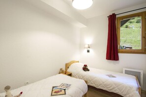 A bedroom with two beds, a window with a red curtain, a wall-mounted lamp, and a view of a green landscape. - Chalet in French Alps with Scenic Views (VALMEINIER)