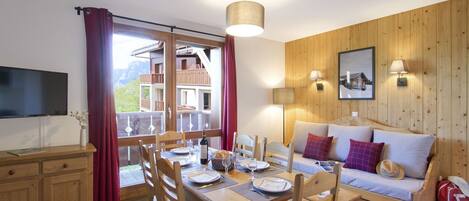 A dining area with a wooden table set for four, a sofa with cushions, a television, and a view of a mountain landscape through a sliding door.
