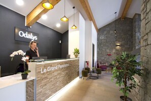 A reception desk with a sign reading 'Le Grand Panorama,' a receptionist, and a stone wall in the background.