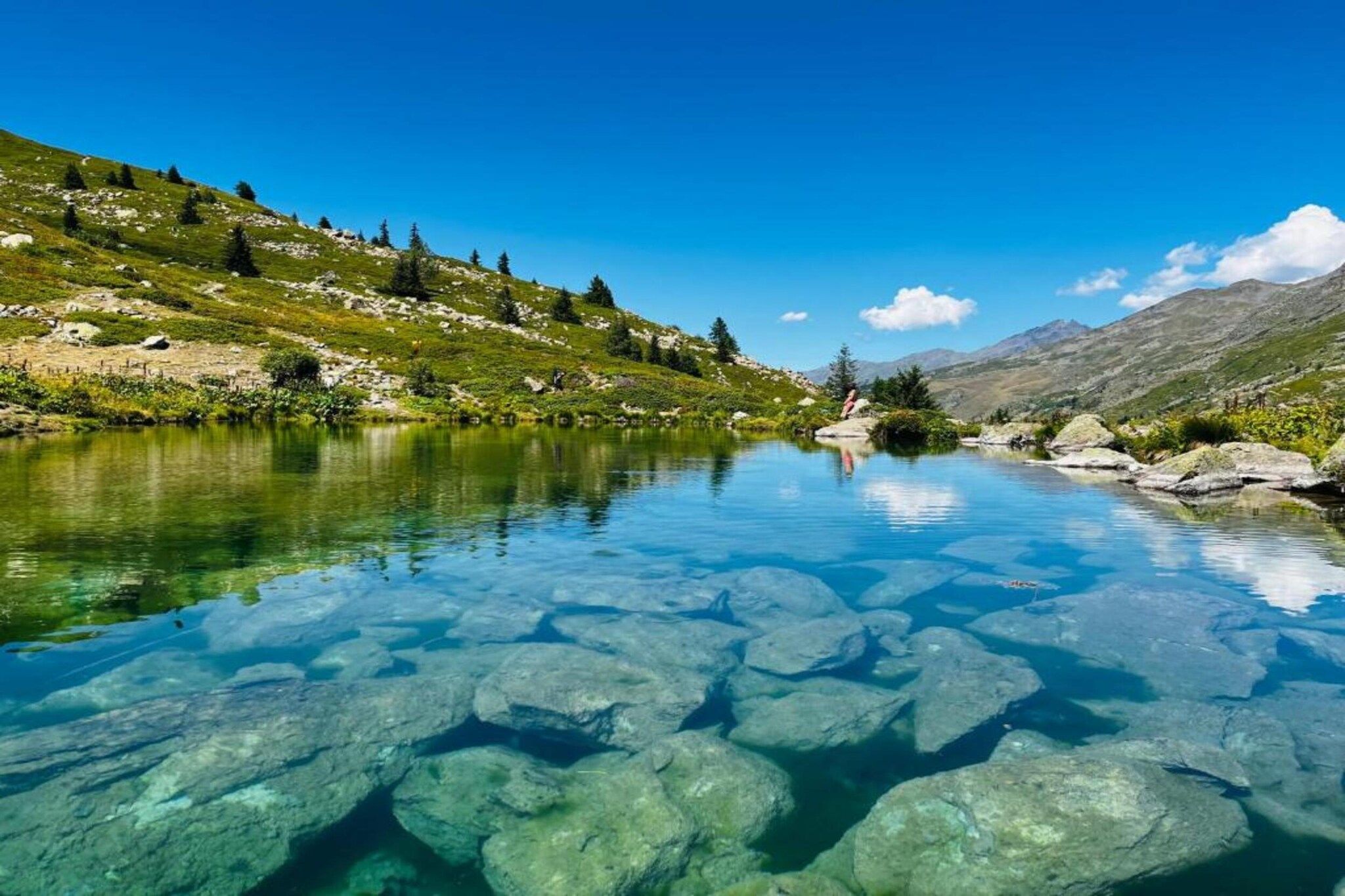 A clear mountain lake with visible rocks and a hilly landscape.