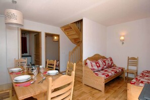 Ein gemütlicher Raum mit Esstisch für vier Personen, einem hölzernen Bett mit gemustertem Überwurf und einer Holztreppe, sowie einer an der Wand befestigten Leuchte.